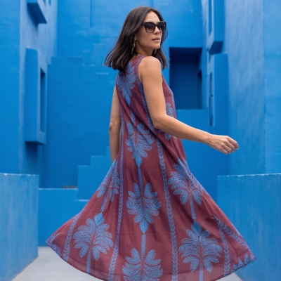 Mulher com vestido midi castanho com padrão azul e óculos escuros em fundo azul arquitetónico.