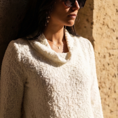 Mulher com casaco branco texturizado e óculos de sol escuros encostada numa parede de pedra