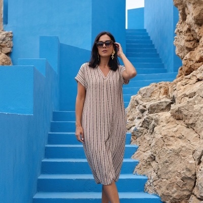 Mulher com vestido midi bege e padrão castanho, sandálias e óculos escuros em escadas azuis com rochas ao lado