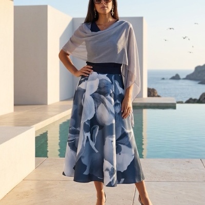 Mulher com vestido azul floral e xale cinza numa varanda com piscina e mar ao fundo.