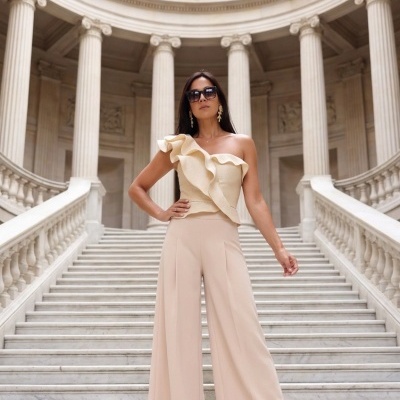 Mulher com blusa bege de um ombro só e calças pantalona bege numa escadaria de mármore com colunas clássicas ao fundo.