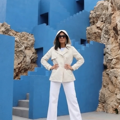 Mulher de casaco bege com capuz e calças brancas junto a paredes e escadas azuis e pedras