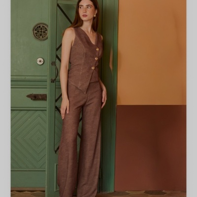 Mulher com conjunto castanho em tecido texturizado, colete com botões dourados e calças largas, sapatos nude, junto a porta verde e parede laranja e castanha