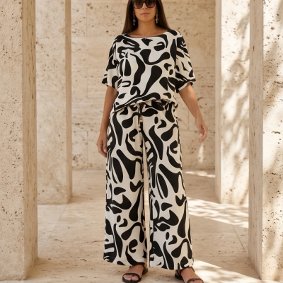 Mulher vestindo conjunto preto e branco com padrão abstrato, blusa de manga curta e calças largas, sandálias pretas e óculos escuros em ambiente com paredes de pedra clara.