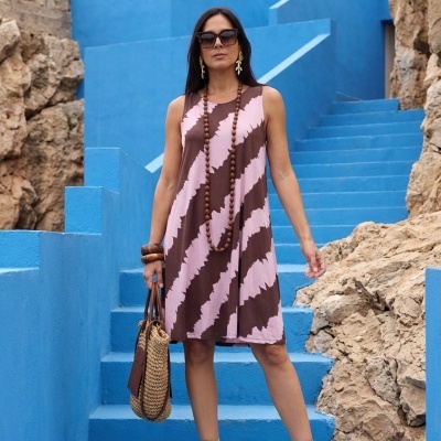 Mulher a usar vestido rosa e castanho com colar e pulseiras castanhas, sandálias douradas, óculos escuros e bolsa de palha em escadas e paredes azuis