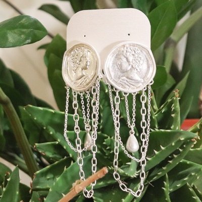 Brincos prateados com medalhões clássicos e correntes penduradas em cartão branco sobre plantas verdes