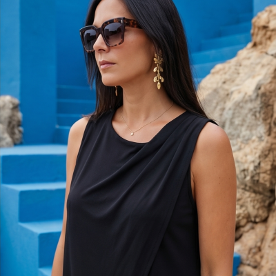 Mulher com óculos de sol padrão tartaruga e vestido preto sem mangas, brincos dourados, com fundo azul e rochas.
