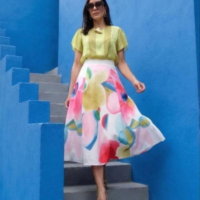 Mulher com blusa amarela, saia branca com flores coloridas e óculos de sol sob escadaria azul