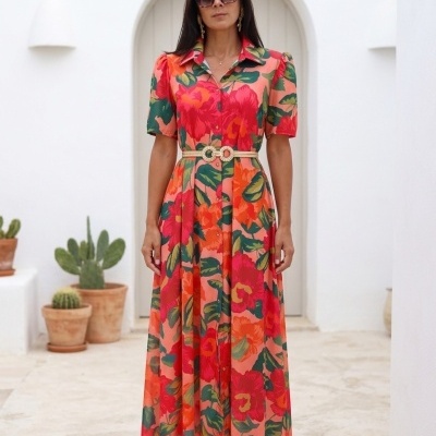 Mulher com vestido longo floral colorido, sandálias douradas e óculos escuros em fundo branco com cactos.