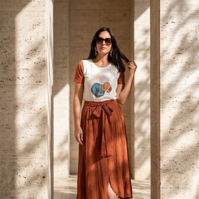 Mulher com roupa casual posando entre colunas de pedra clara em ambiente iluminado pelo sol.