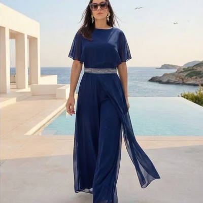 Mulher usando macacão azul-marinho elegante com cinto de pedrarias junto à piscina e vista para o mar.