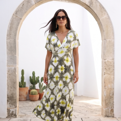 Vestido tie-dye verde, amarelo e branco com decote em V e manga curta, mulher de óculos escuros e brincos, arco de pedra ao fundo.