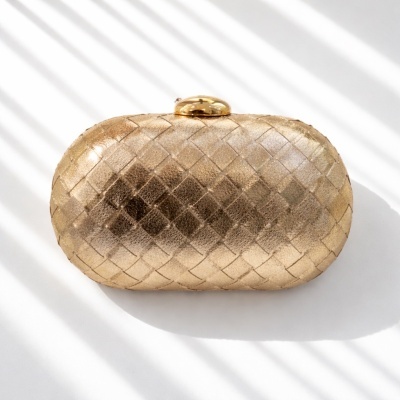Clutch oval dourada trançada com fecho metálico sobre superfície branca