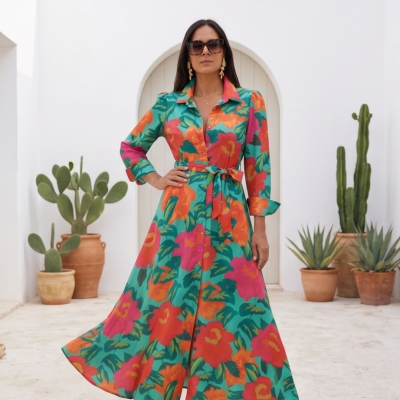 Vestido comprido floral colorido com cinto e botões, usado com sandálias e óculos em ambiente exterior com plantas.
