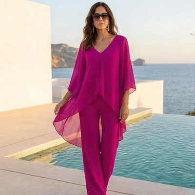 Modelo feminina veste conjunto fúcsia com blusa chiffon e calças largas junto a piscina com mar ao fundo.