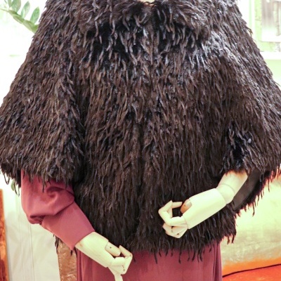 Capa curta castanha de pêlo frisado sobre vestido cor de vinho num manequim.