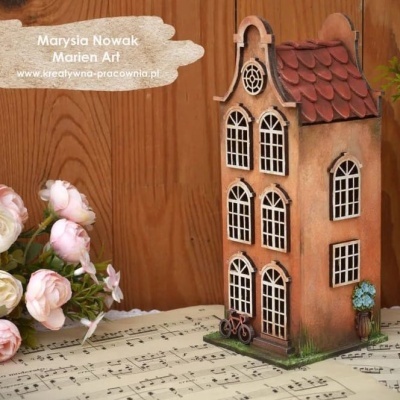 Casa decorativa castanha com janelas brancas e telhado vermelho, com bicicleta e flores ao lado