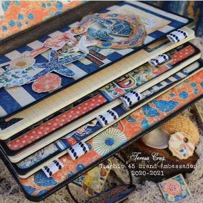 Cadernos com capas decorativas coloridas empilhados sobre tecido e acessórios artesanais.