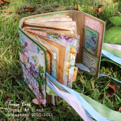 Álbum artesanal decorado com flores, borboletas e laços, sobre relva.