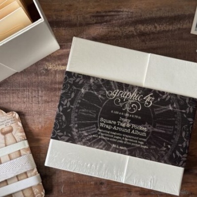 Caderno Square Tag Album branco com faixa preta e textos decorativos sobre mesa de madeira com fitas e fotografias