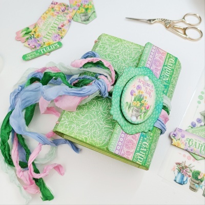 Caderno decorado com padrões e elástico colorido, rodeado por acessórios de papel e uma tesoura dourada.