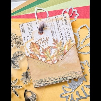 Envelope artesanal com cartão decorativo de folhas de outono e texto de agradecimento num fundo colorido com carimbos e moldes de folhas.