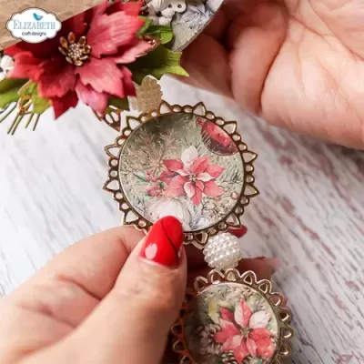 Colar com pendentes redondos florais em metal dourado segurado por mãos com unhas vermelhas.