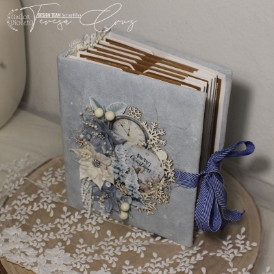 Álbum de fotos com capa cinzenta decorada com flores e laço azul sobre mesa redonda com toalha de renda