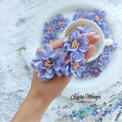 Flor decorativa lilás em seda sobre mão com outras flores num prato branco e chávena