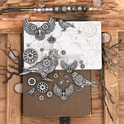 Cartões e recortes metálicos de pássaros e engrenagens sobre livro castanho em madeira com galhos secos.