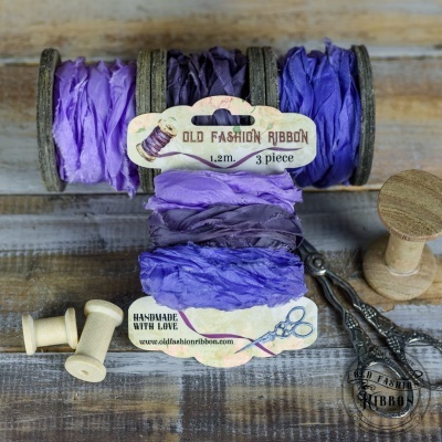 Três fitas de tecido roxas enroladas em carretéis de madeira com etiquetas, ao lado de tesouras antigas sobre mesa de madeira