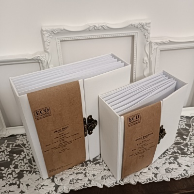 Dois álbuns fotográficos brancos com etiquetas ecológicas e fechos metálicos em cima de tecido rendado com molduras ao fundo.