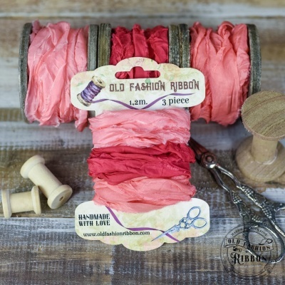 Três fitas de tecido rosa e vermelho enroladas em rolos de madeira com etiquetas e carretéis de madeira ao redor