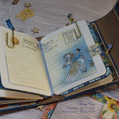 Agenda ou caderno forrado em tecido com páginas ilustradas e clipes metálicos aberto sobre mesa de madeira e adesivos coloridos