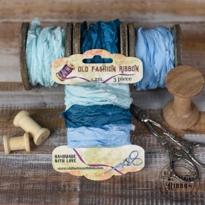 Três rolos de fita de tecido em tons de azul com etiquetas em mesa de madeira, junto a tesoura e carretéis.