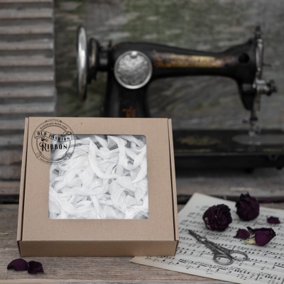 Caixa de fitas brancas em cartão com janela, máquina de costura antiga, partituras, tesoura e flores secas.