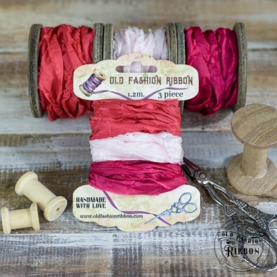 Rolos de fita de tecido embroidery em vermelho, rosa claro e vermelho escuro com etiqueta de papel sobre mesa de madeira clara, com carretéis e tesoura ao lado.