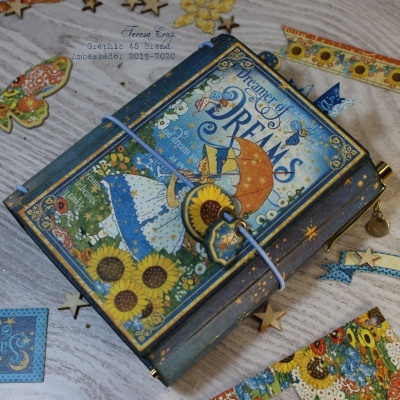 Caderno decorado com capa colorida de girassóis, menina e texto em inglês, fechado com elástico azul sobre superfície de madeira clara com recortes decorativos