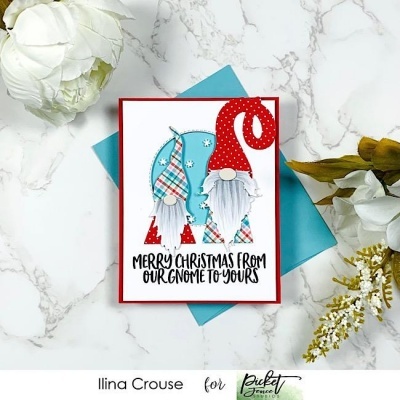 Cartão de Natal com dois gnomos e texto festivo em envelope azul, rodeado por flores sobre superfície de mármore