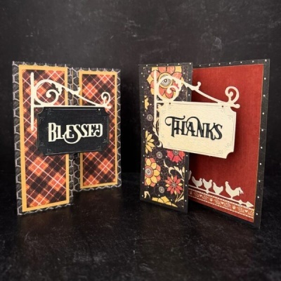 Duas cartas decorativas com textos 'BLESSED' e 'THANKS', padrões xadrez e floral e elementos pendurados em cantoneira branca.