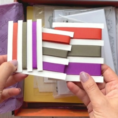 Mãos segurando cartão com fitas coloridas sobre fundo de caixas e papéis