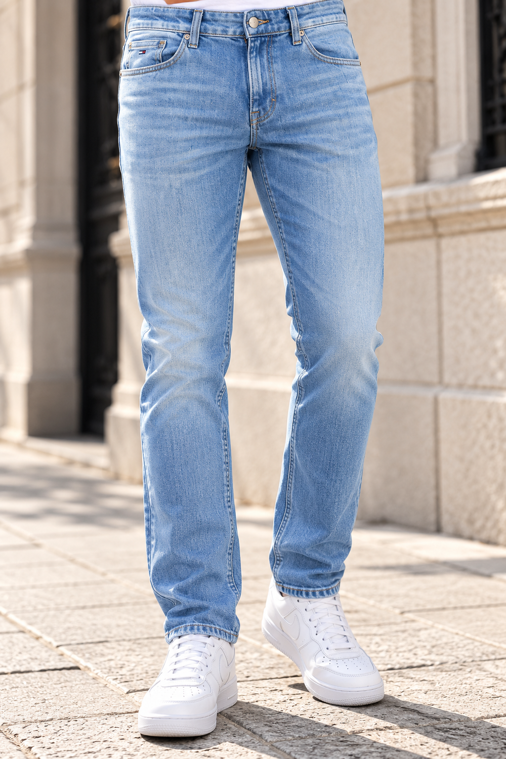 Tommy Hilfiger Austin Slim Tapered Jeans i Lys Blå Calças de ganga azuis claras usadas com ténis brancos na rua junto a uma parede de pedra.