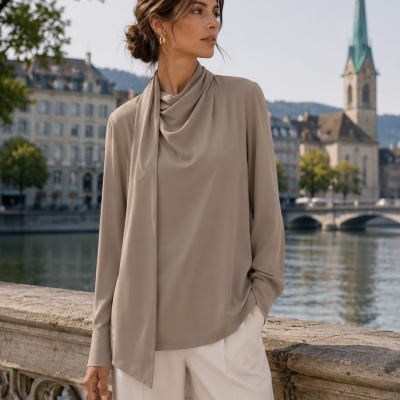Woman in taupe silky blouse and white trousers near stone railing by river with buildings and church steeple in background