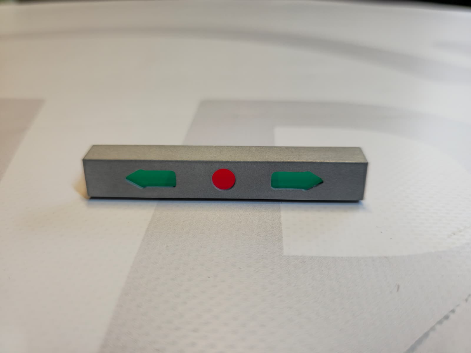 Metallic rectangular object with green arrows and red circle on a white patterned surface