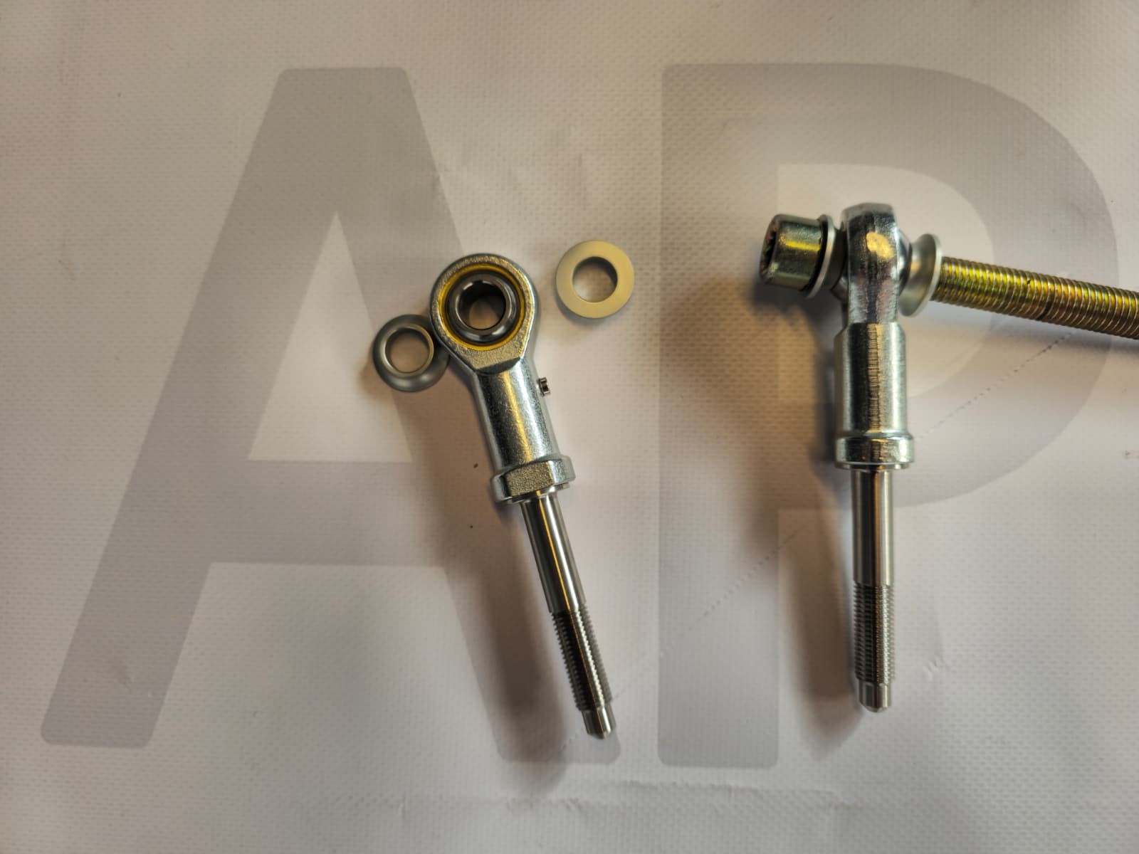Two metal rod ends with spherical bearings and threaded shafts on a white surface with A P text background