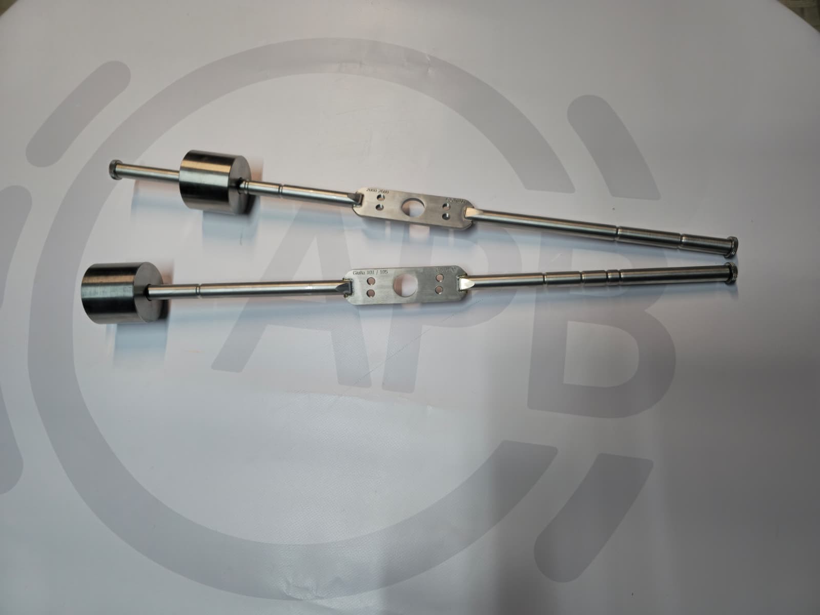 Two metal tools with cylindrical weights and flat sections with holes on a white surface.