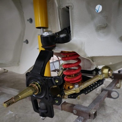 Suspensão automóvel com mola vermelha, amortecedor amarelo, braço preto e componentes metálicos numa estrutura branca