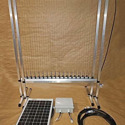Dispositivo metálico com fios, painel solar pequeno, caixa branca e cabos sobre superfície castanha