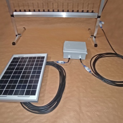 Painel solar retangular com caixa cinzenta e cabos, sobre pano castanho