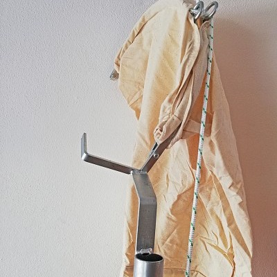 Suporte metálico prateado com tecido bege pendurado e cordão branco com listras verdes junto a uma parede branca.